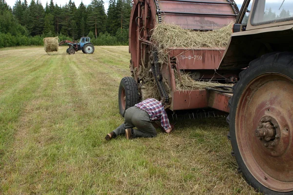 Russian farmer tractor-driver repairs baler hay in the field.