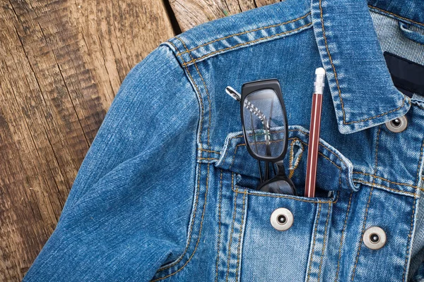 Glasses and pencil in pocket of denim jacket on wooden backgroun