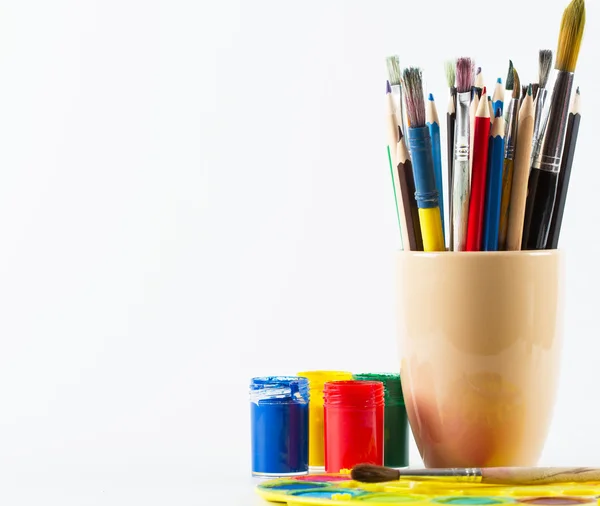 Paint brushes and pencils in mug and colorful paints on white background