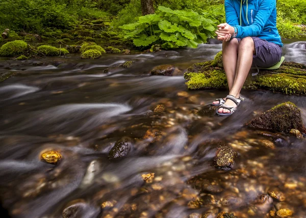 Person sits on the stone covered with moss in the center of rapid flow of the river, holding his feet in clear water