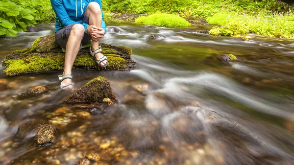 Person sits on the stone covered with moss in the center of rapid flow of the river, holding his feet in clear water