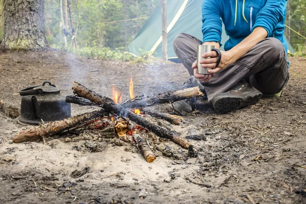 Girl sitting while camping near the fire heated and drink hot tea