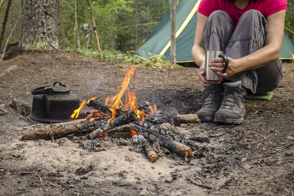 Girl sitting while camping near the fire heated and drink hot tea