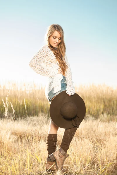 Beautiful young lady model posing in a field at sunrise with a hat in hand