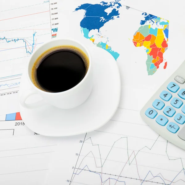 Coffee cup and calculator over world map and stock market charts - close up shot