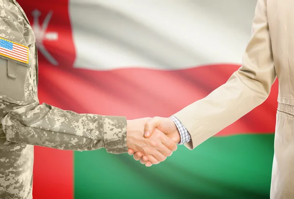 USA military man in uniform and civil man in suit shaking hands with national flag on background - Oman