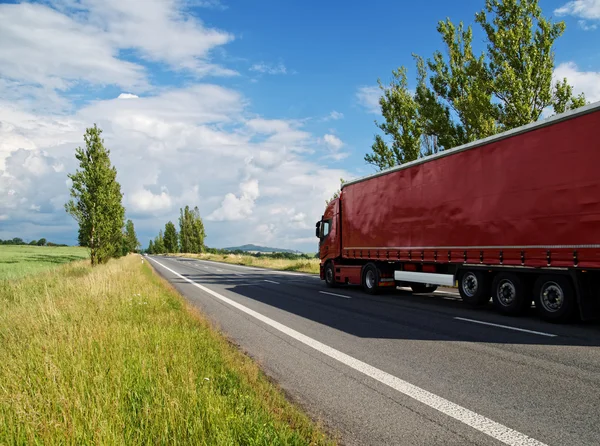 Red truck travels empty road lined with poplar alley in the countryside.