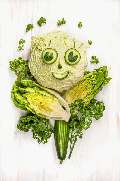 Funny girl face made of  green vegetables, cucumber and lettuce on white wooden background