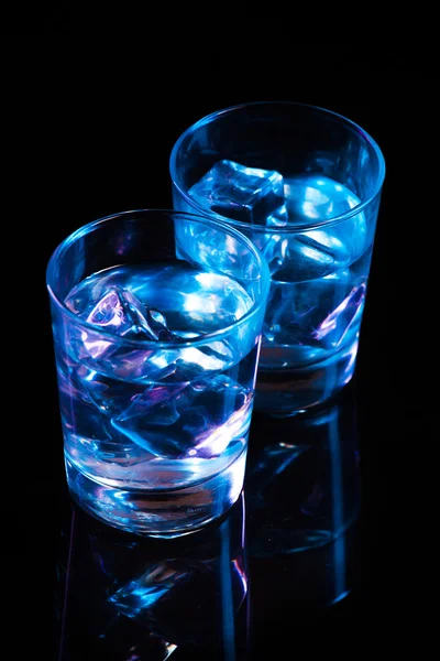 Two glasses with vodka and ice cubes against the background of deep blue glow on dark mirror