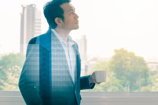 Double exposure of businessman drinking hot coffee by the window with cityscape.