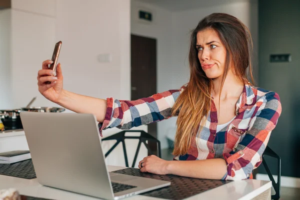 Young girl taking selfie with funny faces in the kitchen. Laptop