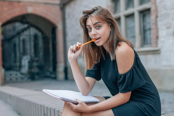 Young student is sitting and holding the pen in mouth. City back