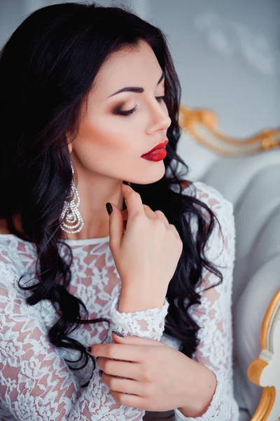 Portrait of perfect, sexy young woman with long black hair and red lips wearing seductive white dress sitting on luxury vintage chair.