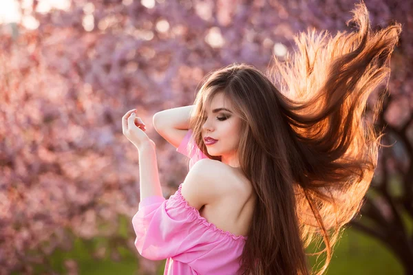Beautiful young woman with long healthy blowing hair running in blossom park at sunset.