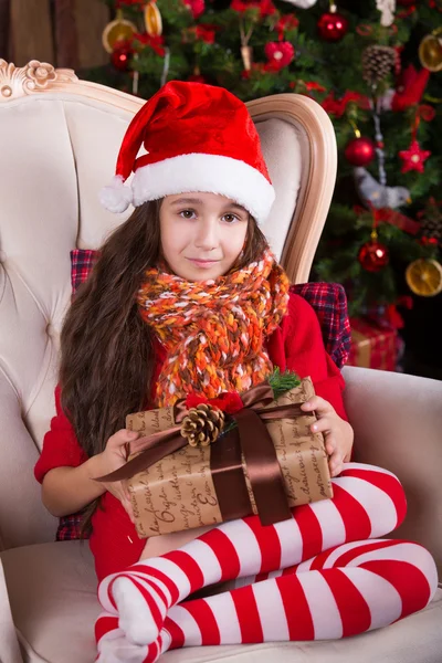 Beautiful Santa girl dreaming near the Christmas tree, holding a present. New year interior