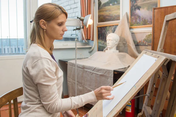 She paints on an easel with a portrait sculpture in the artists studio