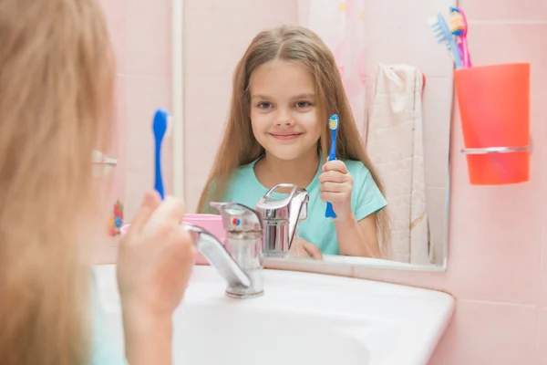 Six year old girl holding a toothbrush and looks at himself in the mirror, while in the bathroom