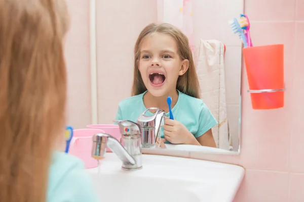 Six year old girl opening her mouth treats teeth in reflection in a mirror, while in the bathroom
