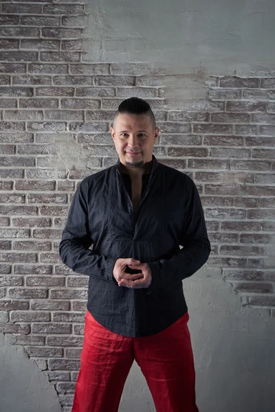 Portrait of a young man, chubby, dressed in red slacks and a black shirt, hands in his pockets, fashionable hairstyle with shaved temples and slicked- back hair at the top of the head, different emotions, Brick wall, fashionable beard