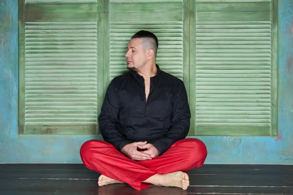 Portrait of a young man, black shirt and red slacks, hairstyle with shaved temples and slicked- back hair at the top of the head, different emotions, green wall, brown floor, beard, lotus posture