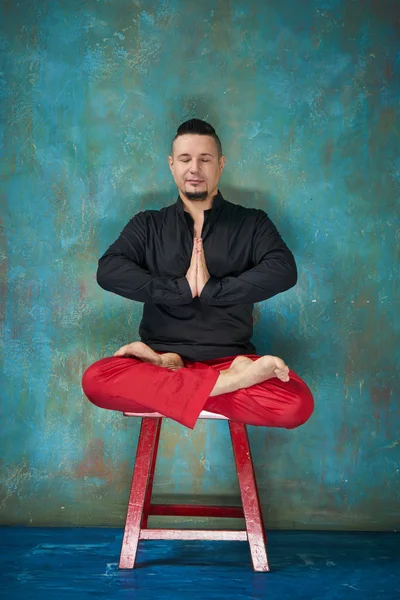 Portrait of a young man, black shirt and red slacks, hairstyle with shaved temples and slicked- back hair at the head, different emotions, green wall, blue floor, beard, lotus posture, red chair