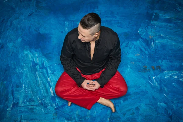 Portrait of a young man, black shirt and red slacks, hairstyle with shaved temples and slicked- back hair at the top of the head, head bowed down, blue floor, beard, lotus posture, hands together