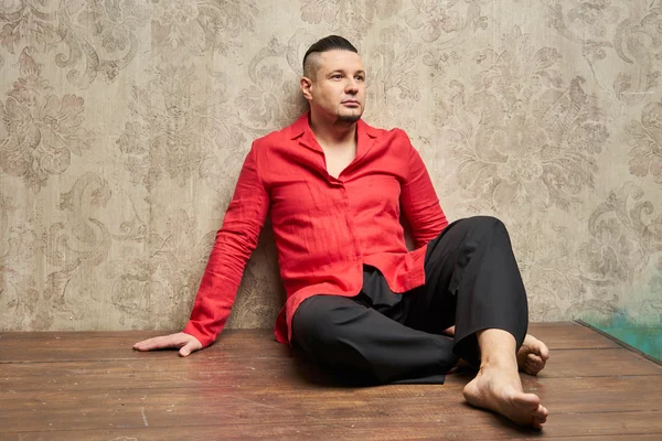 Portrait of a young man, in black slacks and a red shirt, hairstyle with shaved temples and slicked- back hair at the top of the head, emotions, beige wall with drawings, wood brown floor, beard