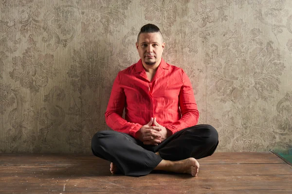 Portrait of a young man, in red shirt and black slacks, hairstyle with shaved temples and slicked back hair at the head, different emotions, wood floor, beard, lotus posture, beige wall with drawings