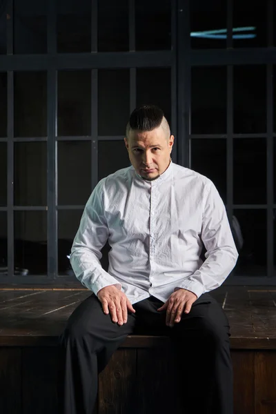 Portrait of a young man, in black slacks and a white shirt, hairstyle with shaved temples and slicked- back hair at the top of the head, different emotions, black window, brown floor, Brick wall, beard, skinhead, brutal, evil, killer