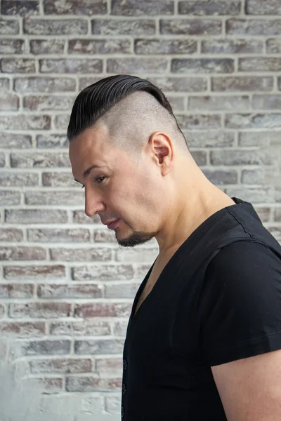 Close-up portrait of a young man, dressed in black T-shirt hairstyle with shaved temples and slicked- back hair at the top of the head, Brick wall, beard, serious face , head bowed down, head profile
