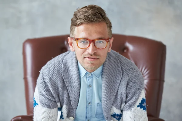 Portrait of a stylish intelligent man with glasses stares into the camera, good view, small unshaven, charismatic, blue shirt, gray sweater, sitting on a brown leather chair