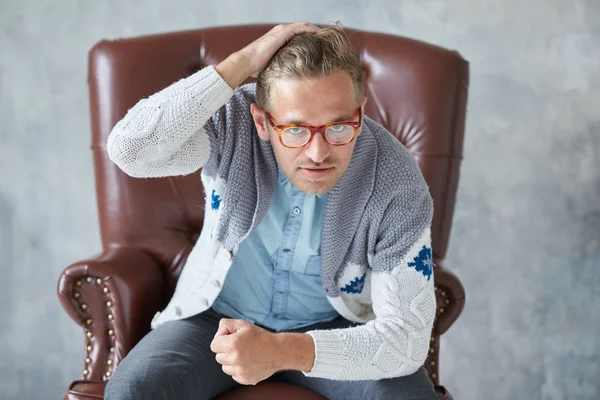 Portrait of a stylish intelligent man with glasses stares into the camera, good view, small unshaven, charismatic, blue shirt, gray sweater, sitting on a brown leather chair, grabs his head