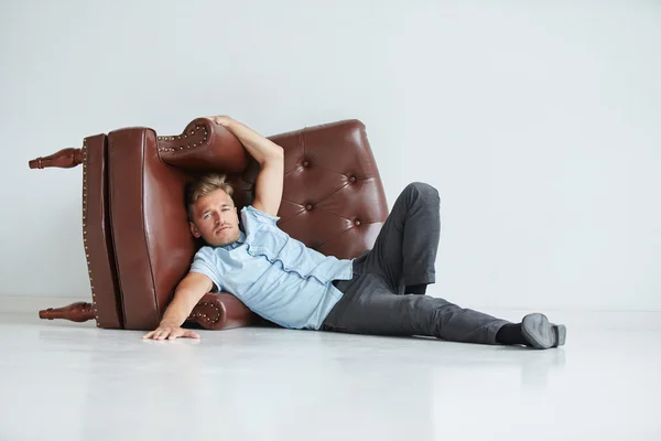 Brutal man lying next to a brown leather armchair , armchair lying on its side on the floor , a man holds hands behind the chair