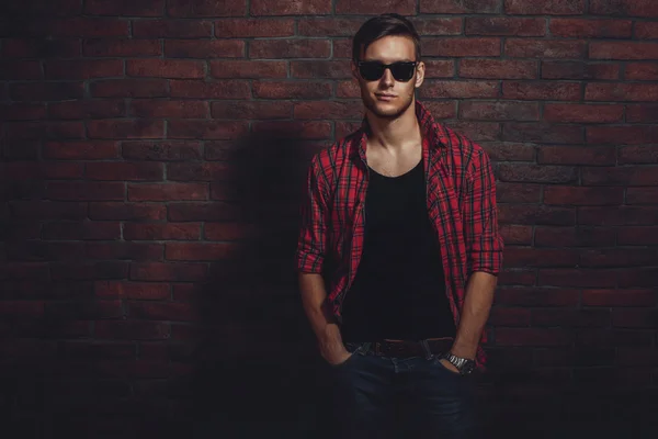 Attractive young hipster smiling and standing against brick wall with hands in the pockets