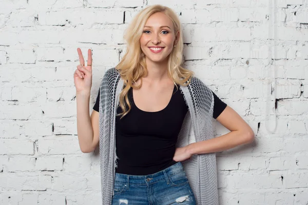 Bright young blond girl is smiling and posing against the brick wall showing v-sign victory or peace with fingers hand gesture