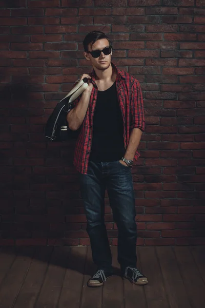 Young hipster man wearing sunglasses casual clothes unbuttoned shirt and denim jeans with bag over shoulder standing near brick wall hand in the pocket looking away