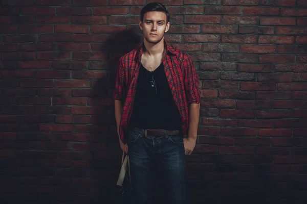 Hipster serious man casual clothes unbuttoned shirt and denim jeans standing near brick wall hand in the pocket looking at camera