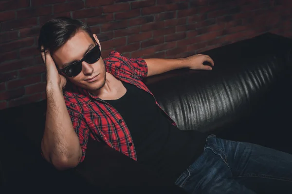 Portrait handsome young hipster man glasses casual clothes unbuttoned shirt sitting relaxing leaning on leather luxury sofa looking away top view