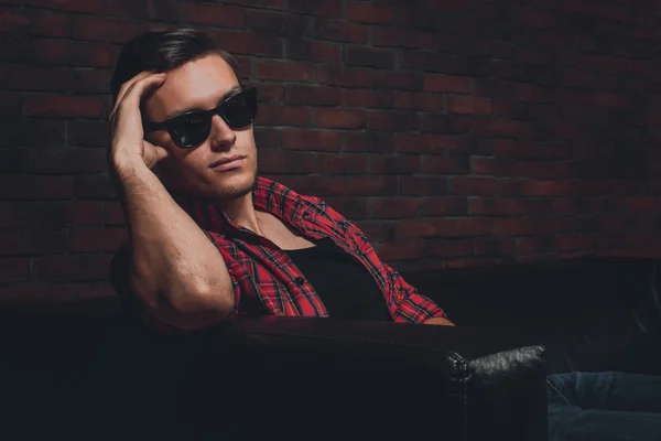Portrait handsome young hipster man glasses casual clothes unbuttoned shirt sitting relaxing leaning on leather luxury sofa looking at the camera