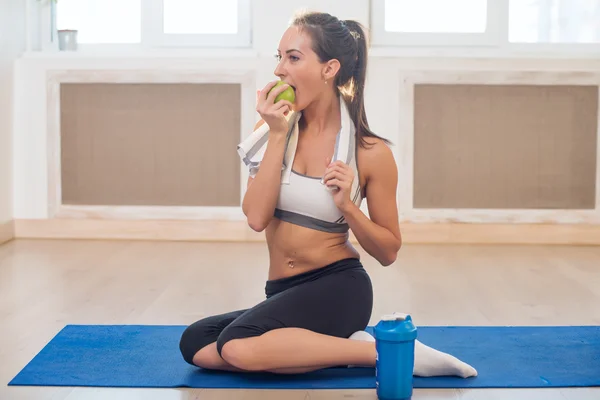 Caucasian beautiful healthy athletic sportive girl eat green apple and Yoga Mat shaker after the training looking at camera.