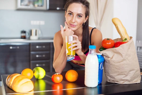 Pretty attractive athletic active sportive lady woman standing in kitchen and healthy food fresh fruits milk bread around holding glass with juice