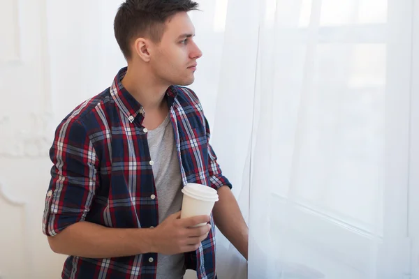 Handsome smiling man guy in plaid shirt looking through the window with a cup of morning coffee concept waiting dreaming