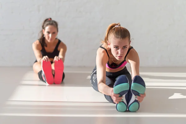 Two young women stretching reaching  with hands to toes seated forward bend warm up at gym fitness, sport, training and lifestyle concept.