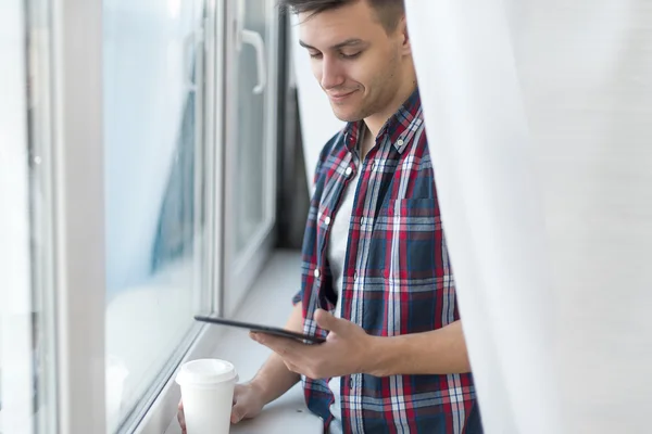 Handsome smiling man guy in plaid shirt near window with a cup of morning coffee and tablet