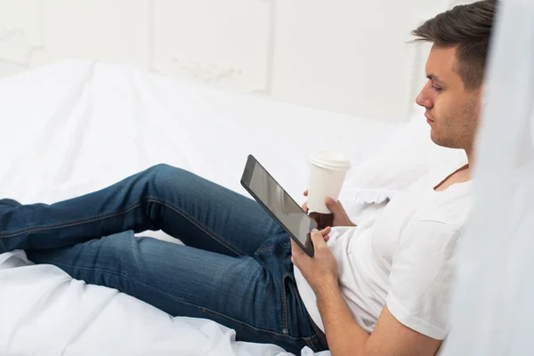 Side view of a casual man in bed at home browsing internet and drinking coffee