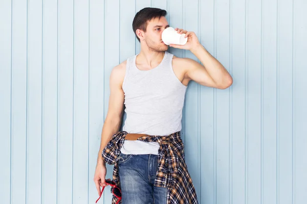Portrait young man holding paper cup and drinking coffee city casual urban style looking at camera standing near wooden wall.