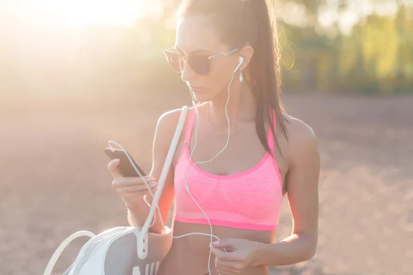 Athlete woman listening music looking at smartphone after workout in nature outdoors portrait summer evening on the beach holidays and vacation healthy lifestyle