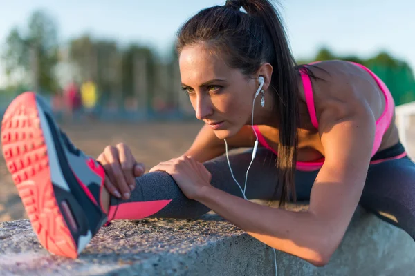 Athletic woman stretching her hamstring, legs exercise training fitness before workout outside on a beach at summer evening with headphones listening music