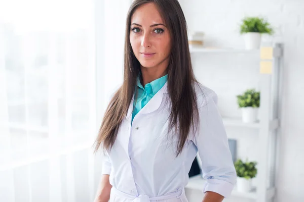 Portrait of young woman doctor with white coat standing in medical office looking at the camera.