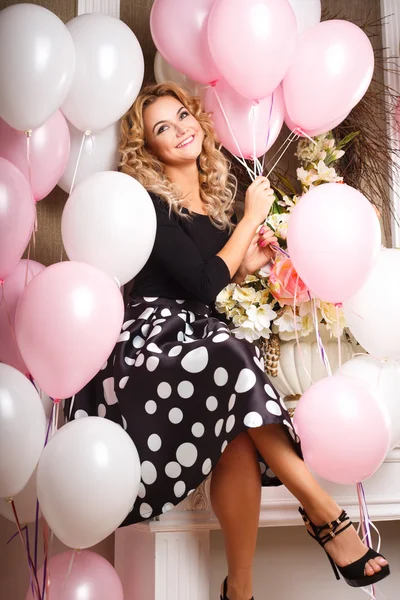 Beautiful curly young woman in a room with many pink and white air balloons. Beauty fashion portrait